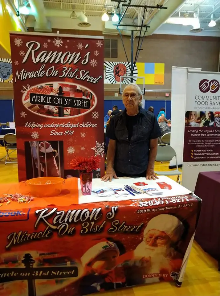 An elderly man stands behind a table at an indoor community event. The table is covered with items and a banner for "Ramon’s Miracle on 31st Street," highlighting holiday support for underprivileged children.