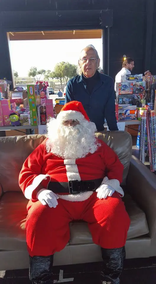 A man dressed as Santa Claus sits on a sofa surrounded by gift-wrapped presents and toys. Another man stands behind him, smiling. The background features a stack of colorful gifts and a partially open door letting in sunlight.