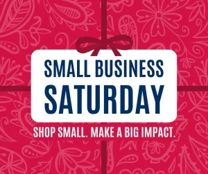 A red background with white floral patterns features a central white box with bold blue text reading "Small Business Saturday." Below in smaller text: "Shop Small. Make a Big Impact." A red bow decorates the top of the box.