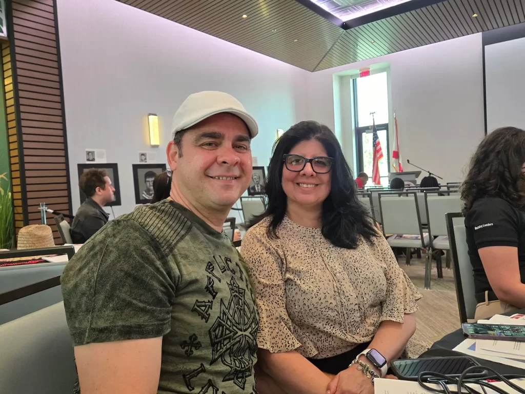 Un hombre con gorra blanca y una mujer con gafas sonríen sentados a la mesa de una sala de conferencias. La sala tiene techos altos, grandes ventanales y varios asistentes visibles al fondo.