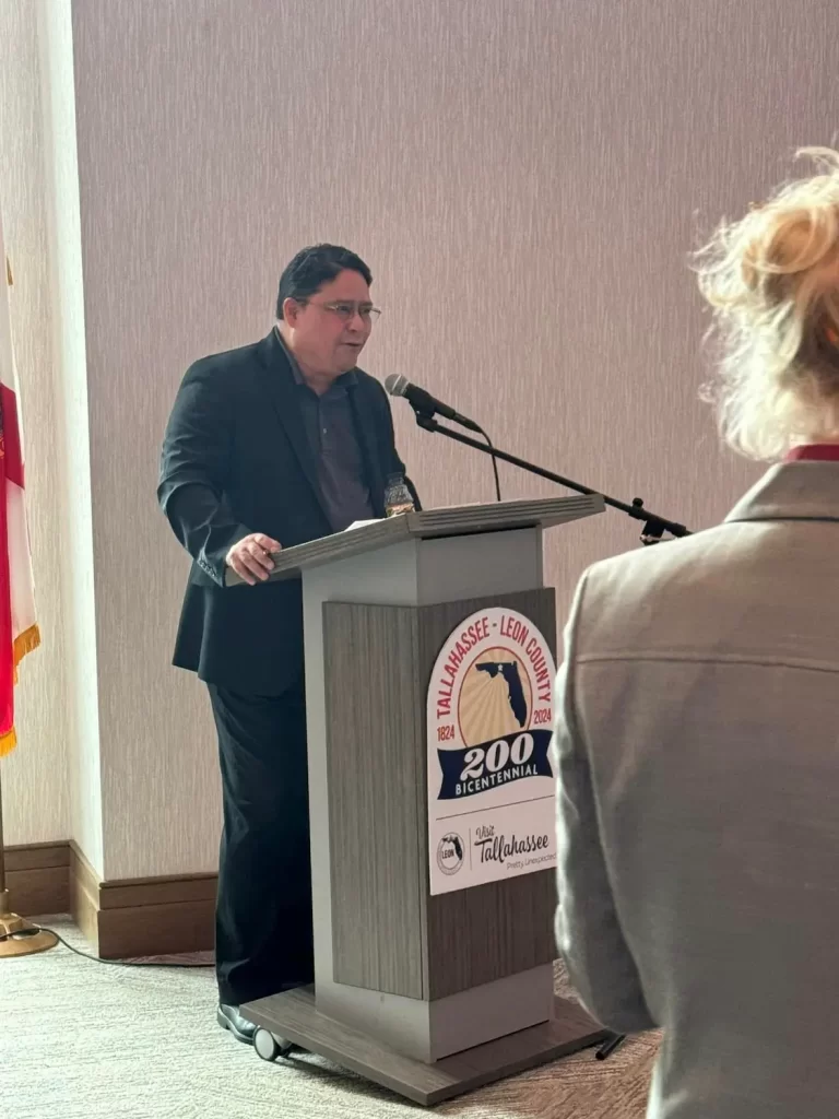 Un hombre está de pie en un podio hablando a través de un micrófono. El podio muestra un logotipo de la celebración del bicentenario del condado de Tallahassee-Leon. Una persona con cabello rubio está en primer plano, parcialmente fuera de foco.