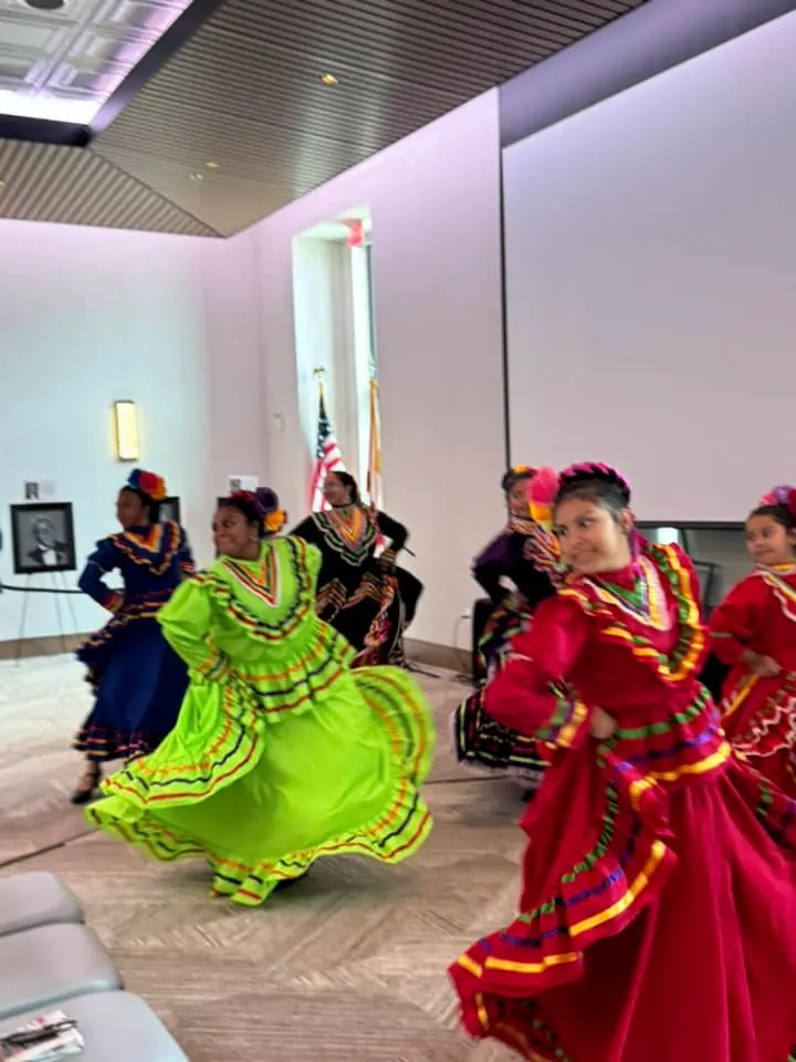 Un grupo de mujeres con coloridos vestidos tradicionales bailan en un espacio cerrado. Los colores vibrantes incluyen verde, rojo y morado, con patrones intrincados. Se ven una bandera estadounidense y un retrato en el fondo.