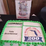 Una torta rectangular decorada con bordes verdes muestra la leyenda "Feliz Bicentenario" y el logotipo del Tallahassee-Leon County 200 Bicentennial. Un cartel detrás dice "Minnie's Cakes" con diseños florales y "Haz lo que te haga feliz y ama".