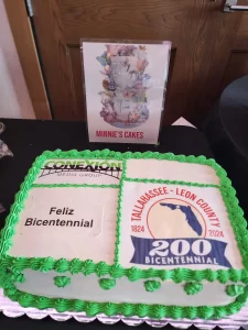 Una torta rectangular decorada con bordes verdes muestra la leyenda "Feliz Bicentenario" y el logotipo del Tallahassee-Leon County 200 Bicentennial. Un cartel detrás dice "Minnie's Cakes" con diseños florales y "Haz lo que te haga feliz y ama".