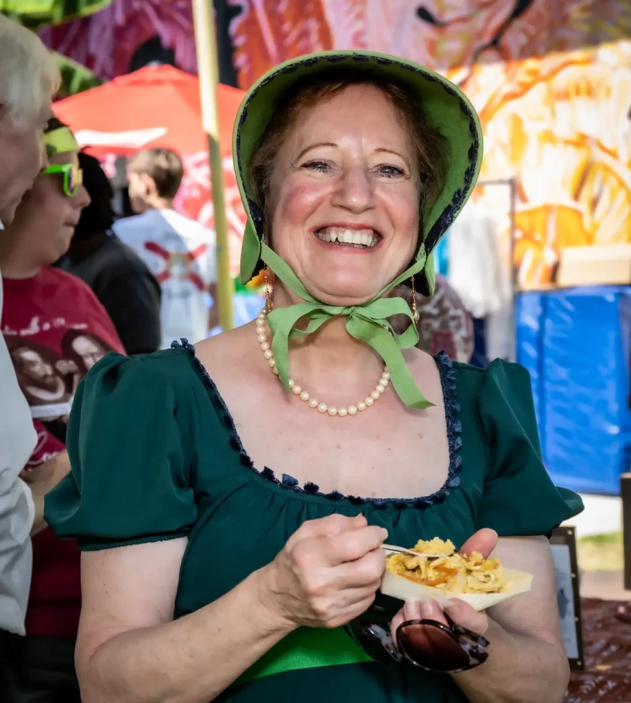 Una mujer sonriente con un sombrero y un vestido verdes sostiene un plato de comida en un evento al aire libre. Lleva un collar de perlas y, al fondo, se ven personas y carpas de colores.