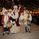 Un grupo de personas vestidas con trajes históricos posa en un animado escenario al aire libre adornado con luces centelleantes. Algunos llevan sombreros y túnicas y se respira un ambiente festivo. Al fondo se puede ver un árbol de Navidad.