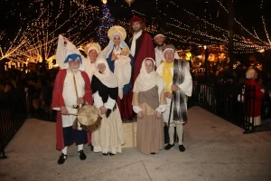 A group of people in historical costumes pose for a photo under festive string lights. They are outdoors, surrounded by a dark night sky, and one holds a drum. Holiday decorations enhance the cheerful atmosphere.