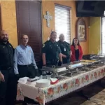 Un grupo de seis personas se encuentra detrás de una mesa de bufé en una sala decorada de forma cálida. La mesa está preparada con bandejas de comida. Una persona se está sirviendo. Las decoraciones de las paredes incluyen una cruz y cuadros.