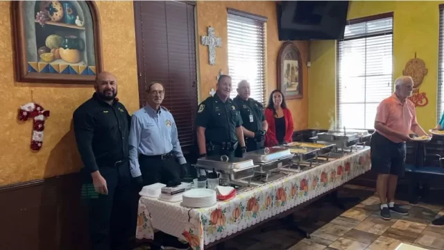 Un grupo de seis personas se encuentra detrás de una mesa de bufé en una sala decorada de forma cálida. La mesa está preparada con bandejas de comida. Una persona se está sirviendo. Las decoraciones de las paredes incluyen una cruz y cuadros.