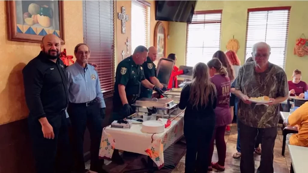 Un grupo de personas sirve comida en una mesa de bufé en una sala con una cálida iluminación. Dos agentes uniformados ayudan, mientras que otros, entre ellos un hombre con camisa azul y otro que sostiene un plato, se encuentran de pie cerca. Se ven mesas de comedor y decoraciones otoñales.