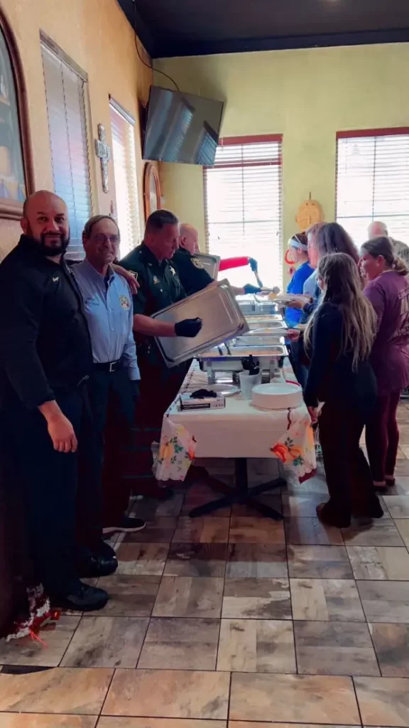 People are standing in line at a buffet table in a restaurant. Some are serving food, while others are waiting. The room has a tiled floor and there are windows in the background.