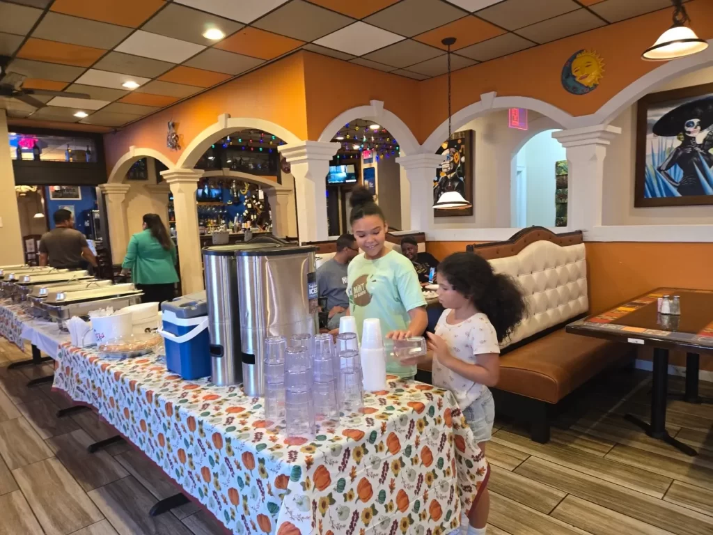 Two children are filling cups at a table covered with a colorful tablecloth in a restaurant. People in the background are near a buffet. The interior has warm tones and themed decor on the walls.