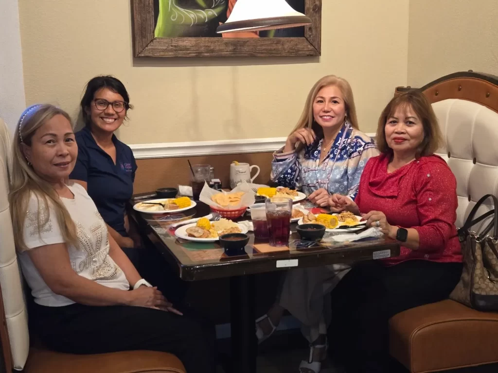 Cuatro mujeres sentadas a la mesa de un restaurante, compartiendo una comida. Sonríen y parecen disfrutar de la compañía mutua. Hay platos de comida y bebidas sobre la mesa y están sentadas en un cómodo reservado.