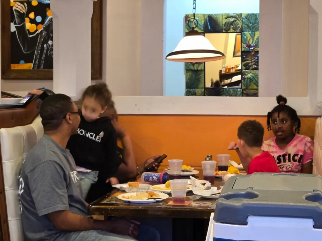 Una familia de cinco personas se sienta alrededor de una mesa de restaurante. Dos adultos y tres niños están comiendo e interactuando. Hay platos y tazas sobre la mesa. El interior es acogedor, con paredes de color naranja brillante y una lámpara colgante. Un espejo refleja la decoración de la habitación.