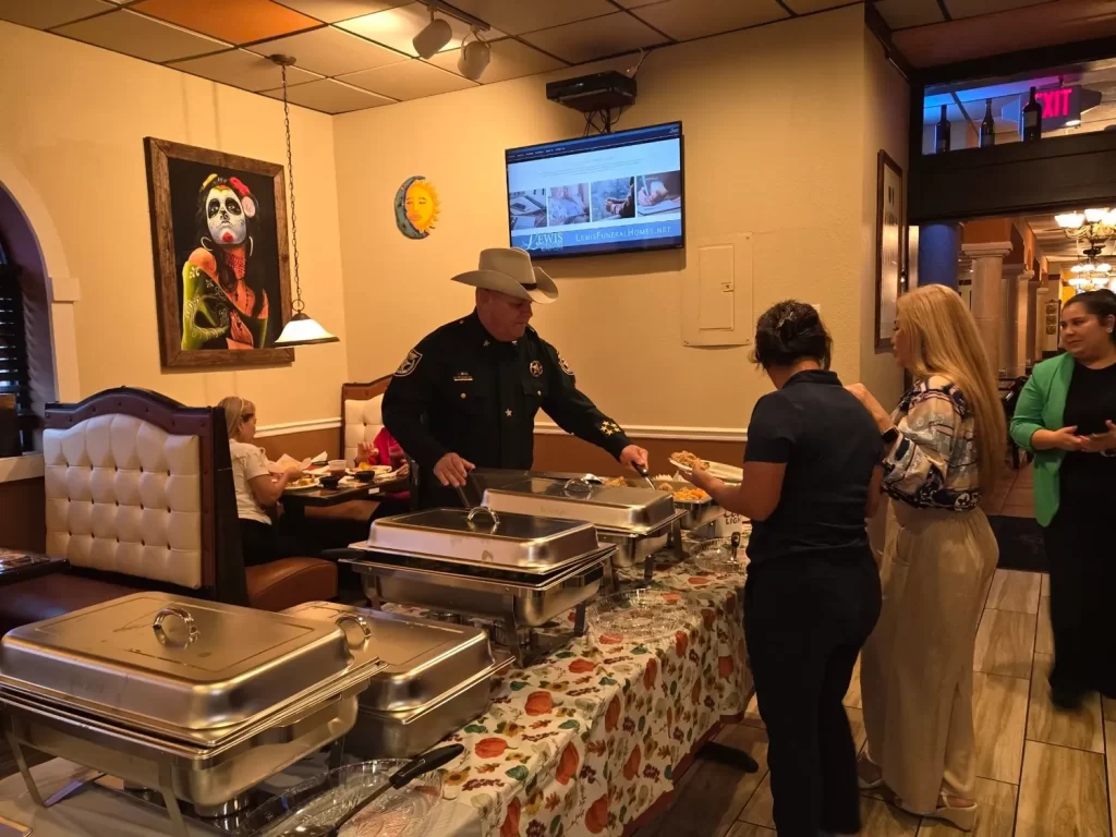 A sheriff in uniform and cowboy hat serves food from a buffet in a restaurant. Two women stand next to him, holding plates. The restaurant features colorful wall art and a table with floral-patterned cloth. Diners are seated in the background.