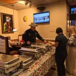 A sheriff in uniform and cowboy hat serves food from a buffet in a restaurant. Two women stand next to him, holding plates. The restaurant features colorful wall art and a table with floral-patterned cloth. Diners are seated in the background.