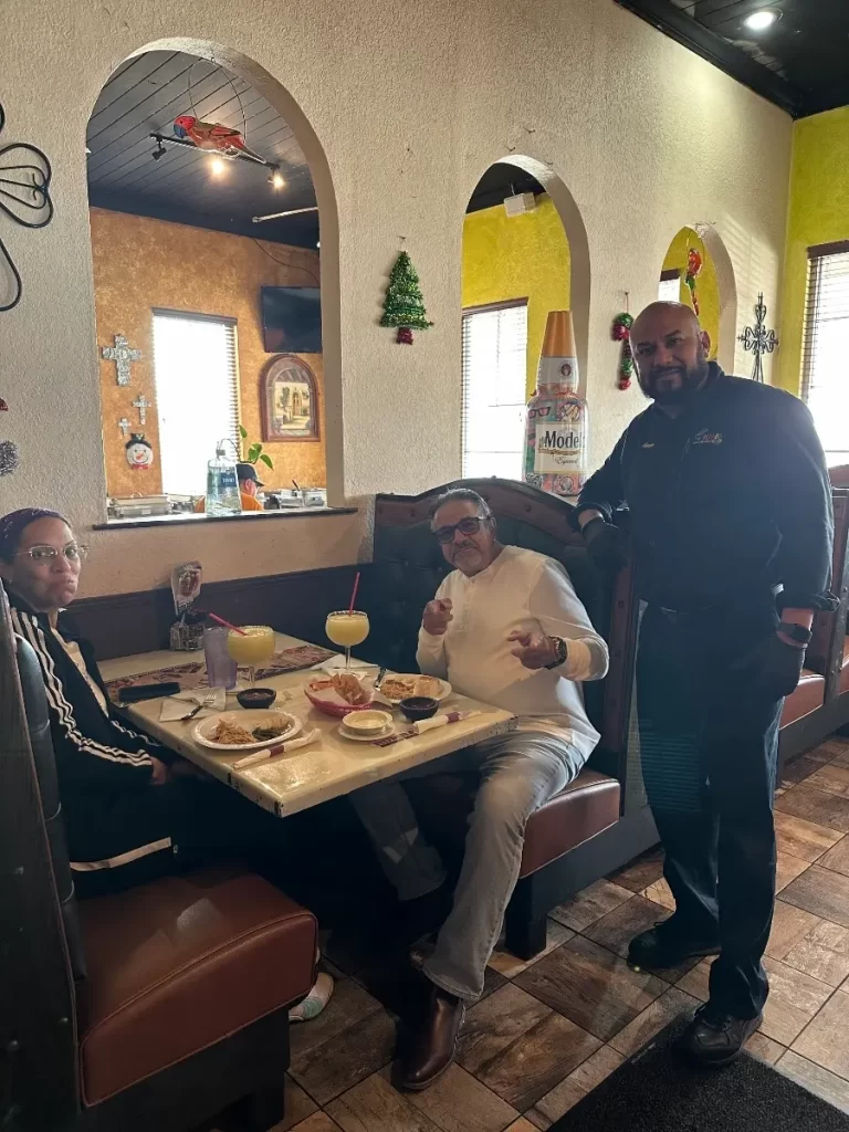 Tres personas en la cabina de un restaurante. Dos están sentadas en una mesa con comida y bebida, y una tercera persona, posiblemente un camarero, está de pie junto a ellas. La decoración incluye paredes coloridas con pequeños adornos navideños.