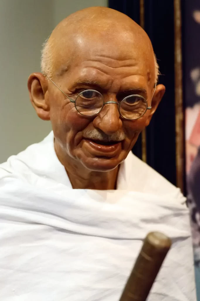 A wax figure of an elderly man with glasses, reminiscent of Gandhi, is displayed in a museum. Wearing a white wrap, the figure has a bald head and gentle expression while holding a wooden staff.