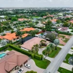 Vista aérea de un barrio suburbano con hileras de casas con techos rojos y marrones, rodeadas de exuberante vegetación y palmeras. Una carretera principal atraviesa la zona y varias casas tienen piscinas en sus patios traseros.