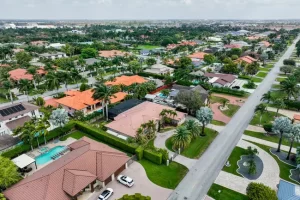 Vista aérea de un barrio suburbano con hileras de casas con techos rojos y marrones, rodeadas de exuberante vegetación y palmeras. Una carretera principal atraviesa la zona y varias casas tienen piscinas en sus patios traseros.