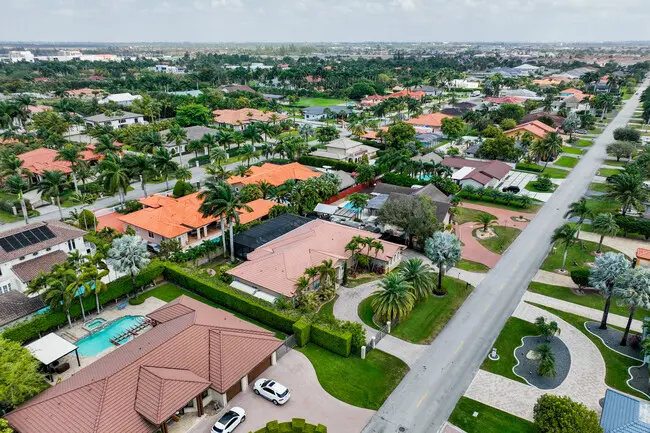 Aerial view of Hialeah, a suburban neighborhood in the United States, featuring rows of houses, palm trees, and swimming pools. The image showcases a long road lined with homes, green lawns, and landscaped gardens against a backdrop of a cloudy sky.