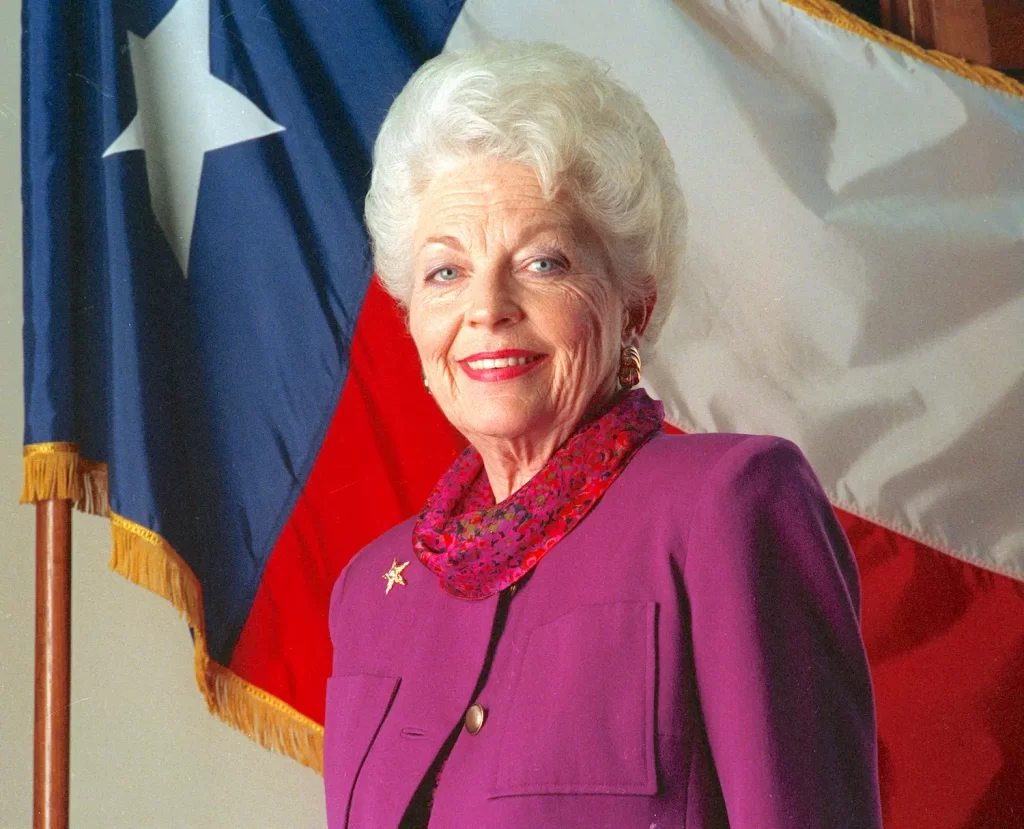 An older woman with white hair smiles while wearing a purple jacket and red scarf. She stands in front of a large flag with a star on it, featuring red, white, and blue colors.