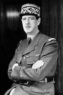 Black and white photo of a uniformed man with a mustache and a patterned military cap. He is standing with his arms crossed. The background is dark and plain.