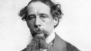 A vintage black and white portrait of an older man with wavy hair, a long mustache, and beard, reminiscent of Charles Dickens. Dressed in a suit, he gazes directly at the camera with a serious expression, evoking the thoughtful demeanor of a renowned author. The background is plain.