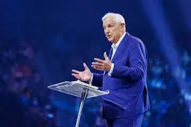 An older man in a blue suit stands behind a clear podium, gesturing with his hands. The background is dark with blurred audience members, suggesting a large event or conference setting. It appears to be an engaging moment, reminiscent of David Jeremiah's distinguished speaking style at major events.