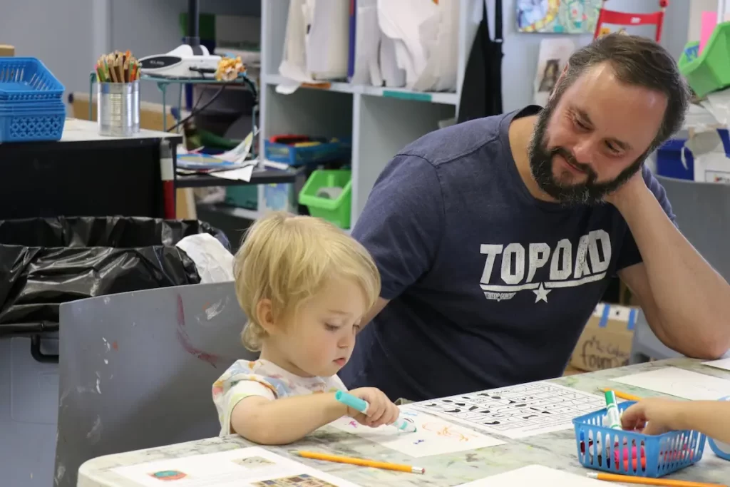 A young child draws with a marker at a table while an adult, wearing a "TOP DAD" T-shirt, looks on smiling. The room resembles a Holiday Light Spectacular with art supplies and children's artwork adding bursts of color to the scene.