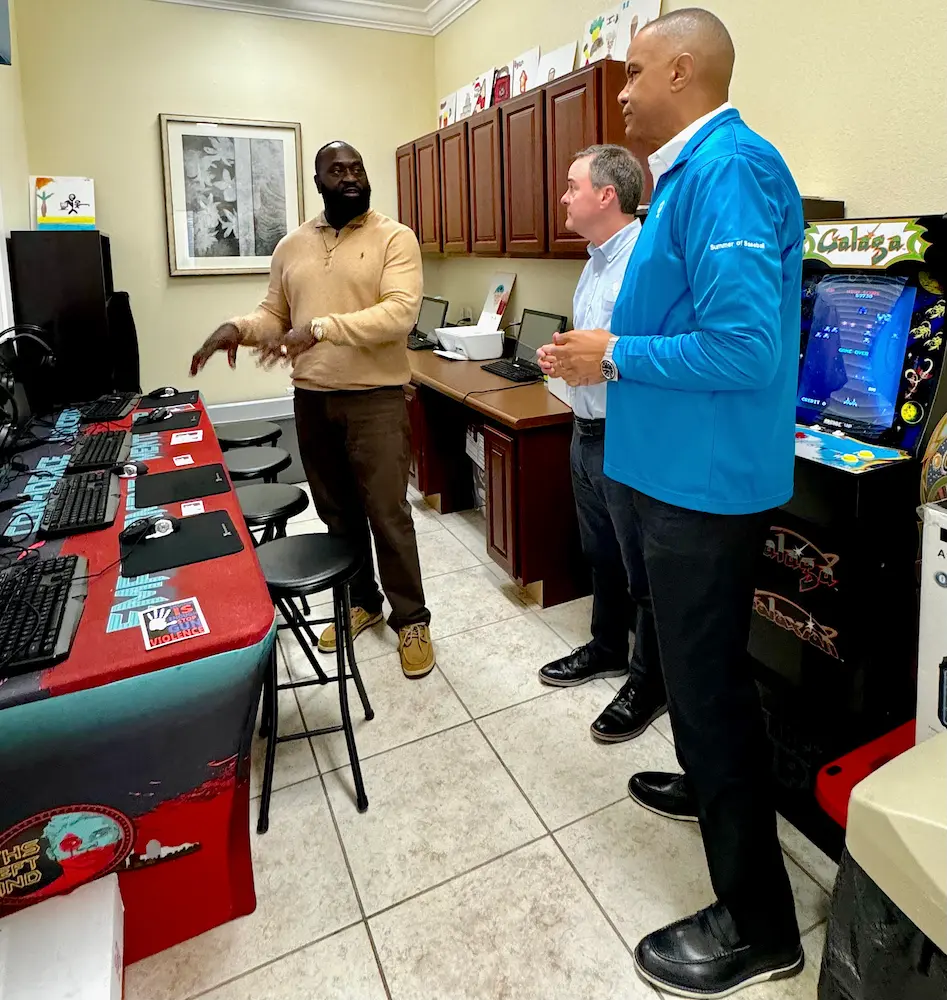 Three men are engaged in conversation in a room with computer stations. One man gestures while the others listen. An arcade machine is in the background, and the floor is tiled.