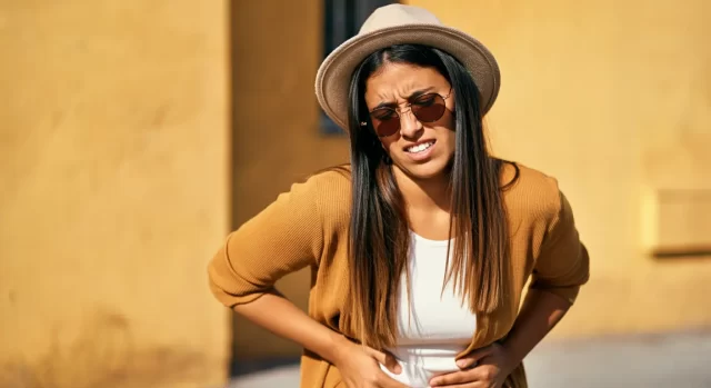 A woman in a brown cardigan and hat stands against a yellow wall, holding her stomach with both hands and expressing discomfort. She wears sunglasses and has long dark hair.