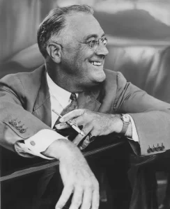 A black and white photo of a smiling man in a suit and tie, leaning on a car door. He wears glasses and holds a cigarette in one hand, appearing relaxed and confident.