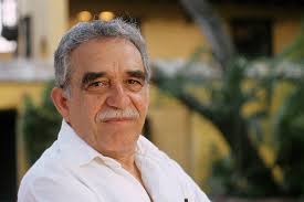 An older man with short gray hair and a mustache is wearing a white shirt. He is outdoors with greenery and a yellow building in the background. He has a neutral expression on his face.