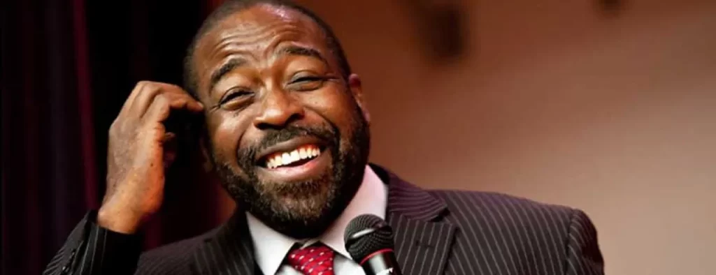 A man in a dark pinstripe suit and red tie smiling broadly while holding a microphone. He appears to be speaking or performing, set against a blurred background.