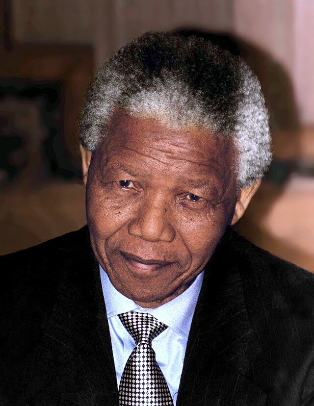 An elderly man with short, curly gray hair and a warm expression is wearing a black suit, white shirt, and patterned tie. The background is blurred and neutral-toned.