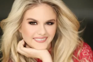 Rachel Barcellona, a woman with long, wavy blonde hair, smiles warmly. She rests her chin on her hand and wears a red top. The blurred background emphasizes her expressive eyes and makeup.