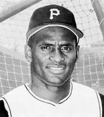 Black and white photo of a baseball player wearing a cap with a "P" on it and a jersey. The background shows part of a baseball field's netting and structure.