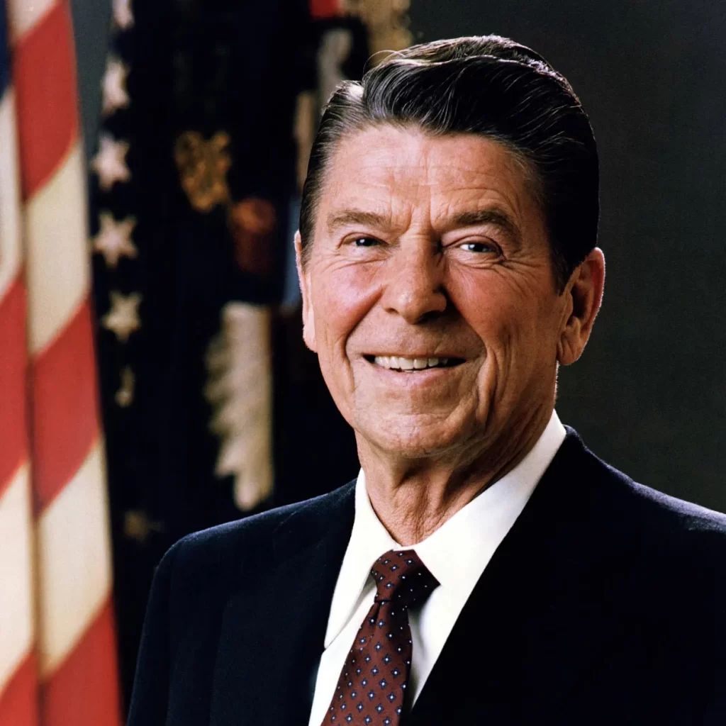 A man in a suit with a white shirt and red polka dot tie stands smiling in front of a U.S. flag and a dark background.