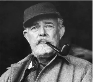 An elderly man with a beard and mustache is wearing a cap and coat. He is smoking a pipe and looking directly at the camera. The background is dark and blurred.