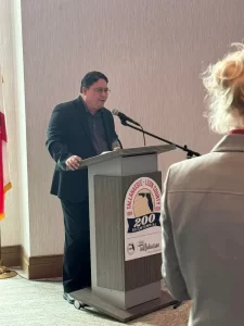 A person stands at a podium with a microphone, speaking. The podium has a "Tallahassee-Leon County 200th Bicentennial" logo. Another person is slightly visible in the foreground, facing away.