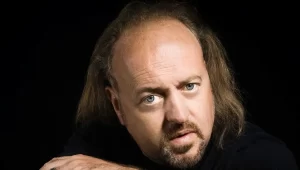 Man with shoulder-length hair and a beard, gazing intently at the camera against a dark background. He wears a black shirt and rests his arms on a surface in front of him.