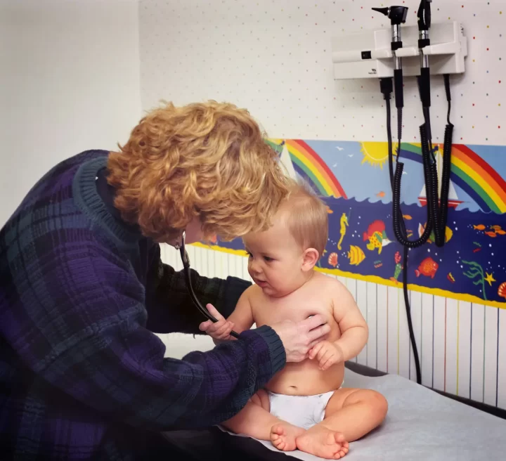 Un profesional de la salud con un suéter violeta examina a un bebé con un estetoscopio en el consultorio de un médico. La habitación tiene decoraciones de pared con motivos de peces y arcoíris de colores.