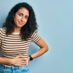 A person with curly hair, wearing a striped shirt and jeans, stands against a light blue background. They have one hand on their hip and a neutral expression.