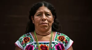 A woman with long dark hair stands against a dark background, wearing a colorful traditional garment adorned with floral patterns and beads. She gazes directly at the camera with a calm expression.