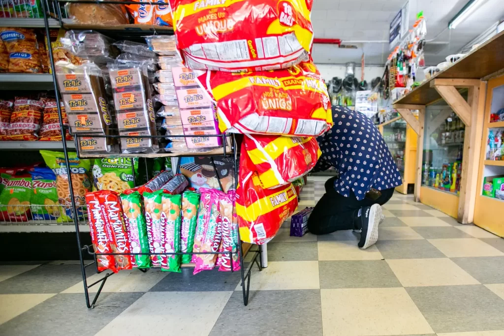 Person kneeling to organize items on a store shelf, surrounded by snacks like chips and pastries. The floor has a black-and-white checkered pattern, and a wooden counter is partially visible on the right.