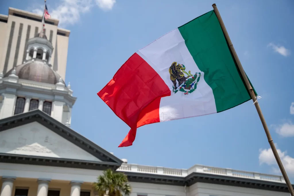 A Mexican flag waving in front of a government building with a dome and columns, under a clear blue sky.