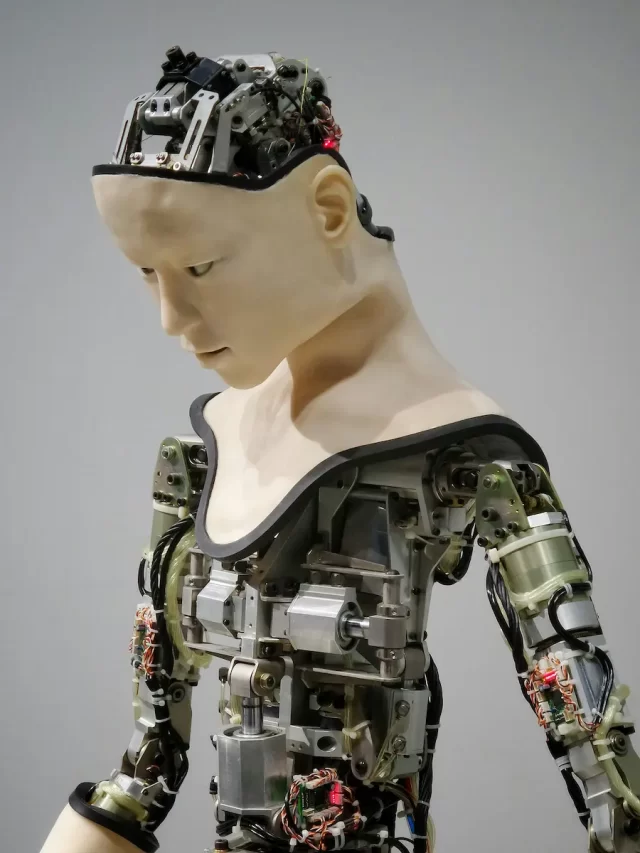 A humanoid robot with exposed mechanical components, dressed in a skin-like covering on the head and shoulders, is captured in a side view against a plain background. Its AI-driven design highlights intricate circuits and metal parts.