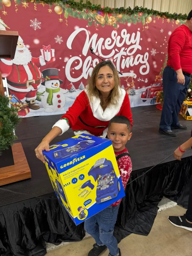 A woman and a child smile as they hold a large Goodyear toy box, standing in front of a festive "Merry Christmas" backdrop with Santa and a snowman. The joyful scene captures the spirit of serving the community during this holiday event.