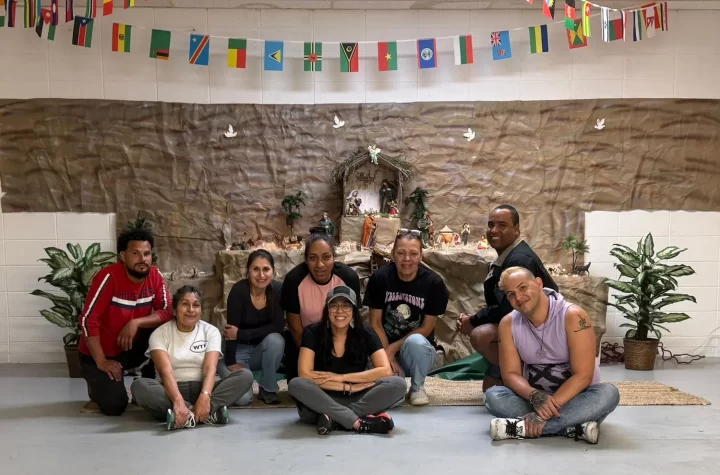 A group of people seated in front of a wall adorned with flags and a festive display reminiscent of the Three Wise Men. They are smiling, casually dressed, and flanked by potted plants, with string lights casting a warm glow.